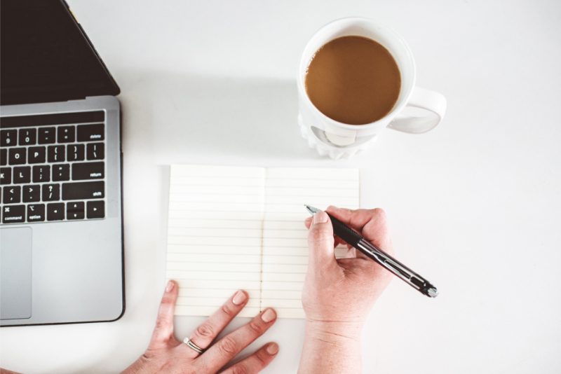 Hands writing in a notebook beside a laptop and coffee, calm and focused workspace